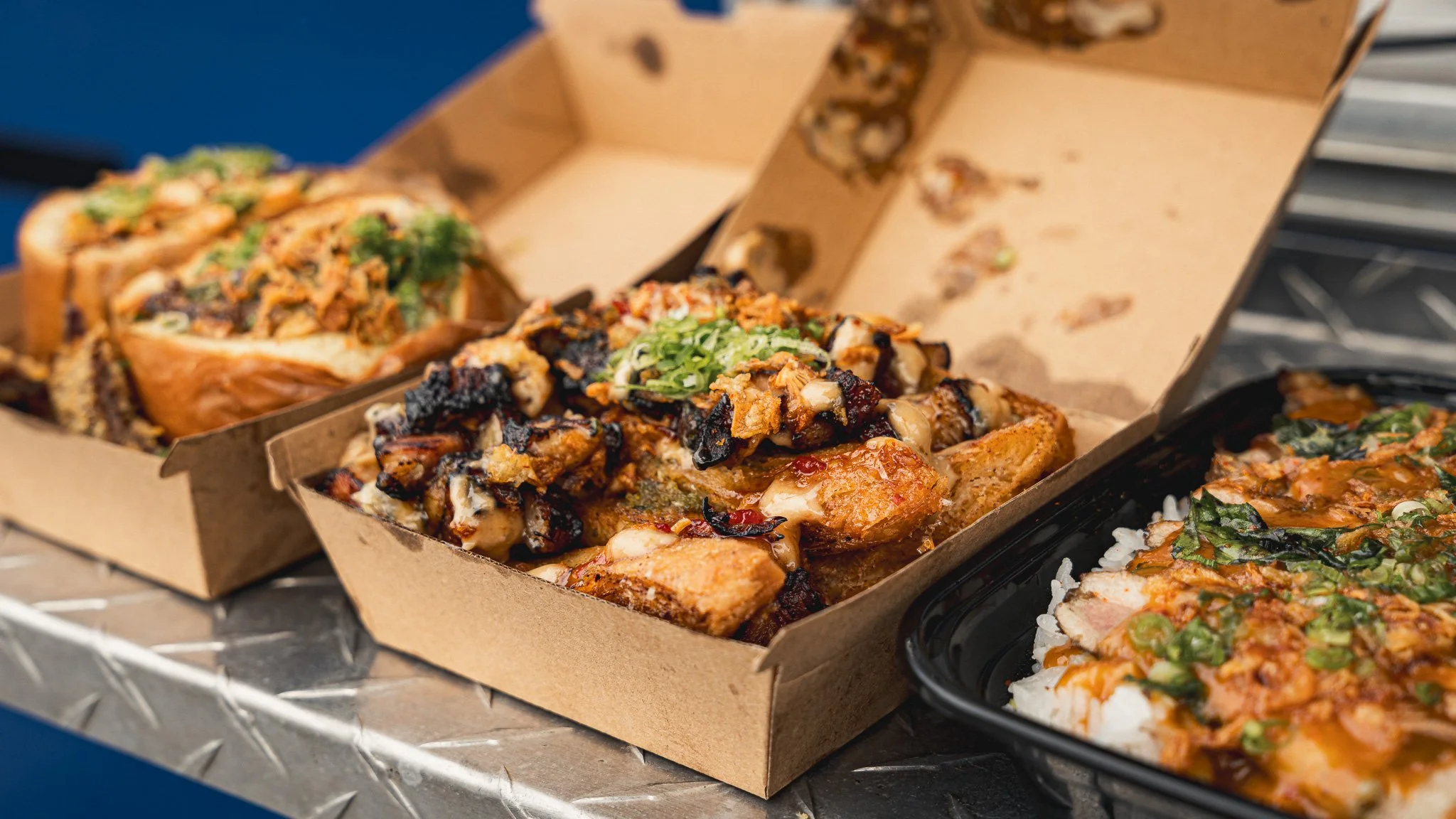 Various types of Asian fusion dishes with grilled meats, rice, and vegetables in takeout boxes.