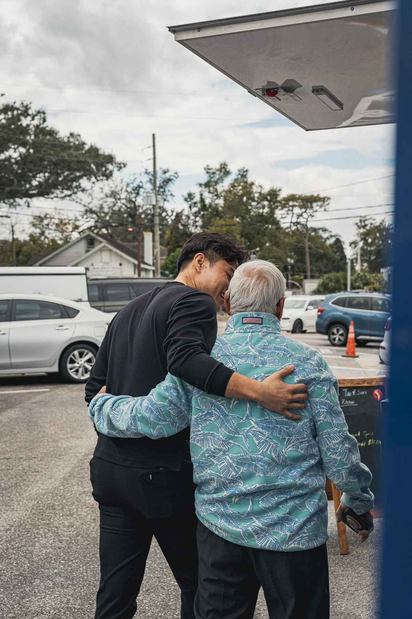 A young man and an elderly man are sharing a joyous hug outdoors in a parking lot, with cars and trees in the background.