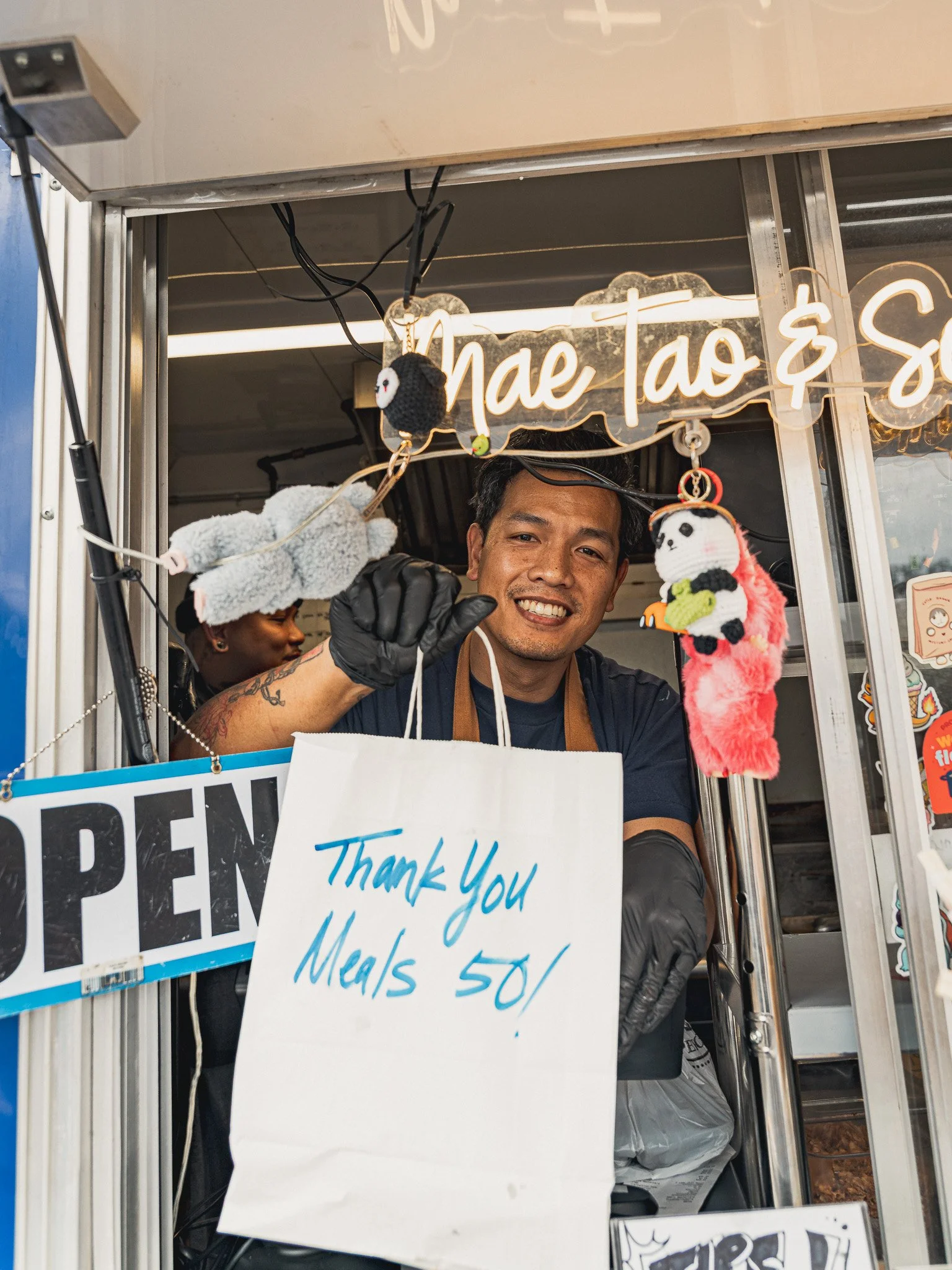 Man smiling inside food stand, holding a plush panda puppet, with signs advertising vegetarian food and meal prices.