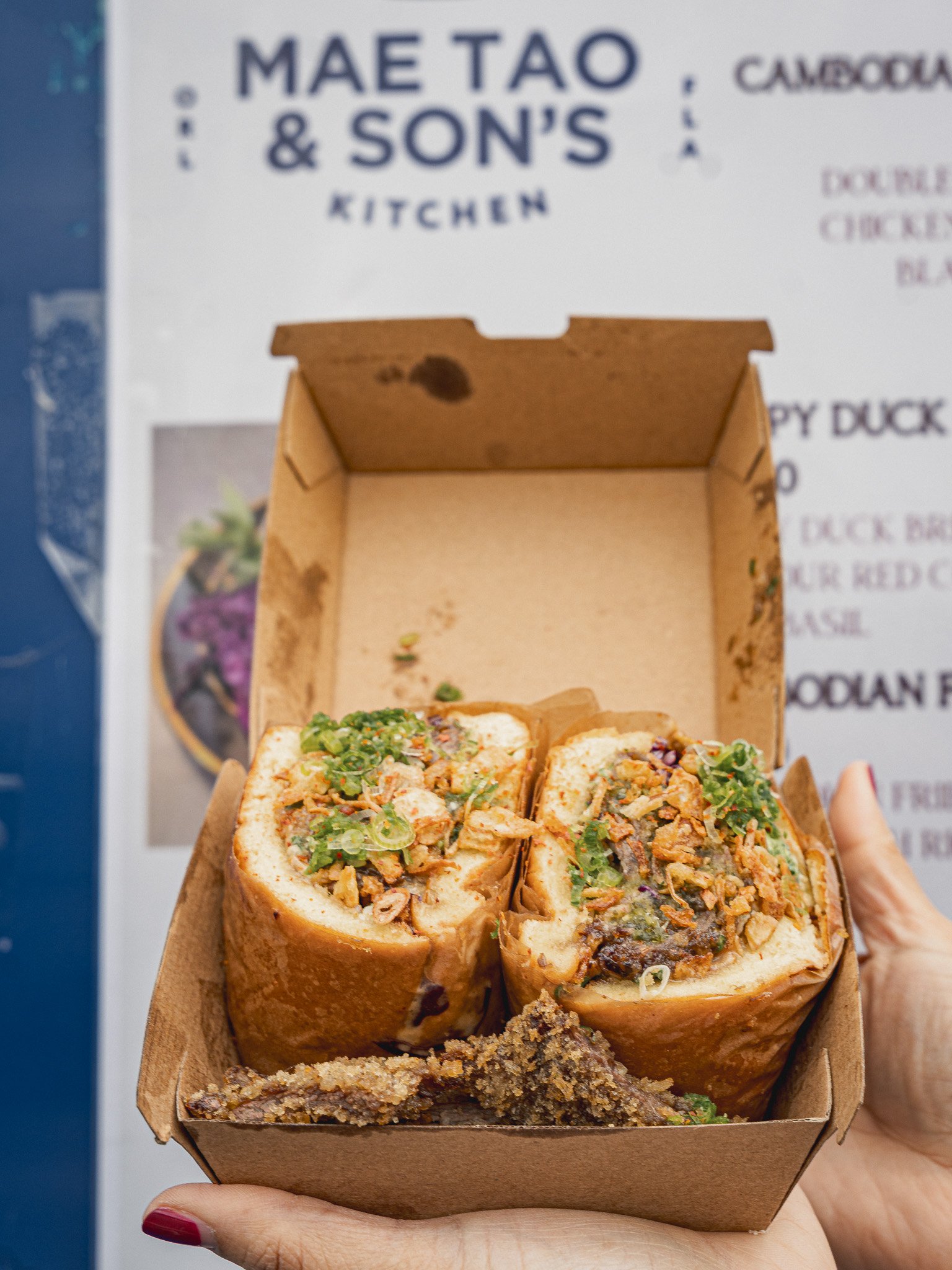 A hand holding a brown takeout box filled with three large sushi burritos with chopped vegetables and sauce. In the background, a sign reads 'MAE TAO & SON'S KITCHEN'.