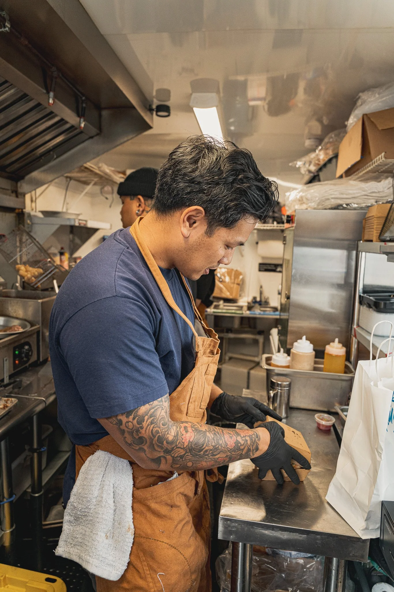 A man with tattoos on his arms, wearing a brown apron and black gloves, is preparing food in a commercial kitchen. He is focused on his task while a woman in the background is also working. The kitchen has stainless steel surfaces, shelves with boxes