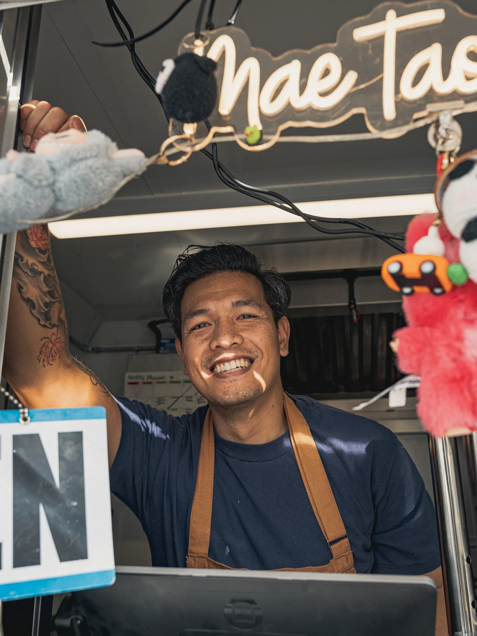 Smiling man with dark hair, tattoos on his arm, wearing a dark shirt and a brown apron, standing behind a counter in a food stall with a illuminated sign above that says 'ne T'.