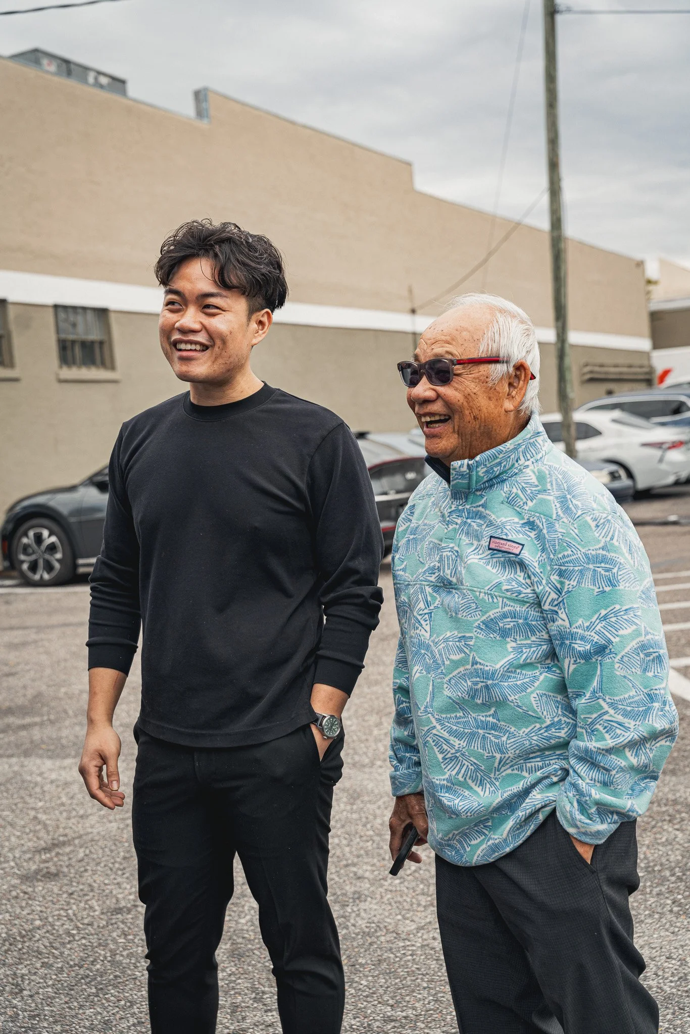 Two men walking outdoors in a parking lot, smiling and talking. One appears to be middle-aged with light gray hair and sunglasses, wearing a patterned shirt. The other is younger, wearing a black long-sleeve shirt, with dark hair, and has his right h