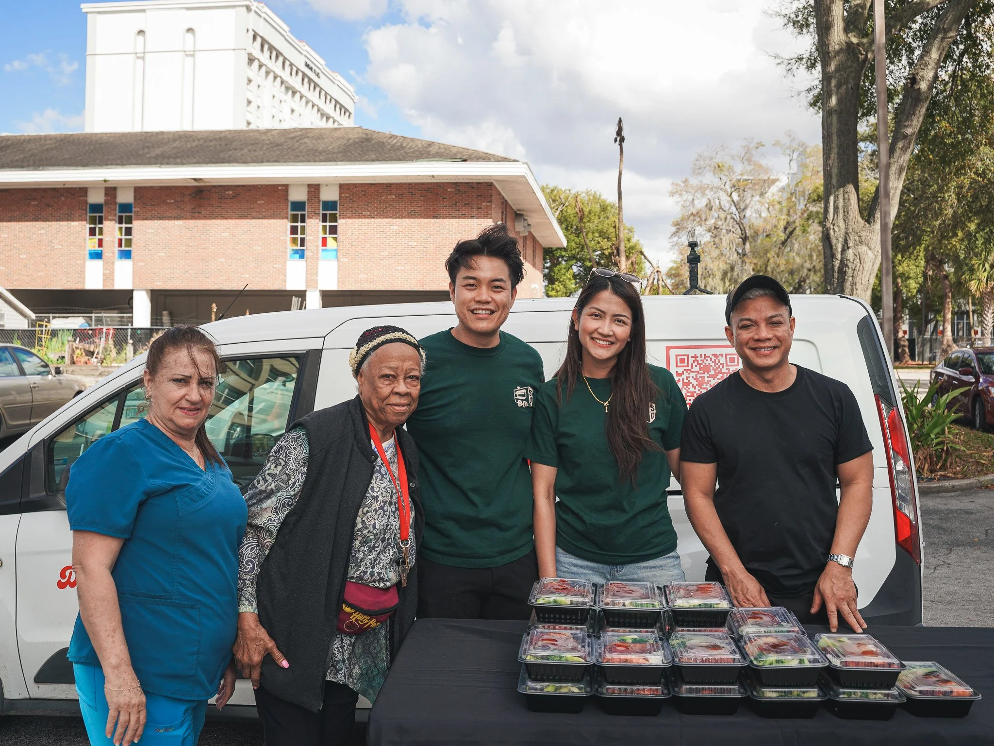 Group of five Meals 50 people standing behind a table with containers of food outdoors, with a white van and buildings in the background.