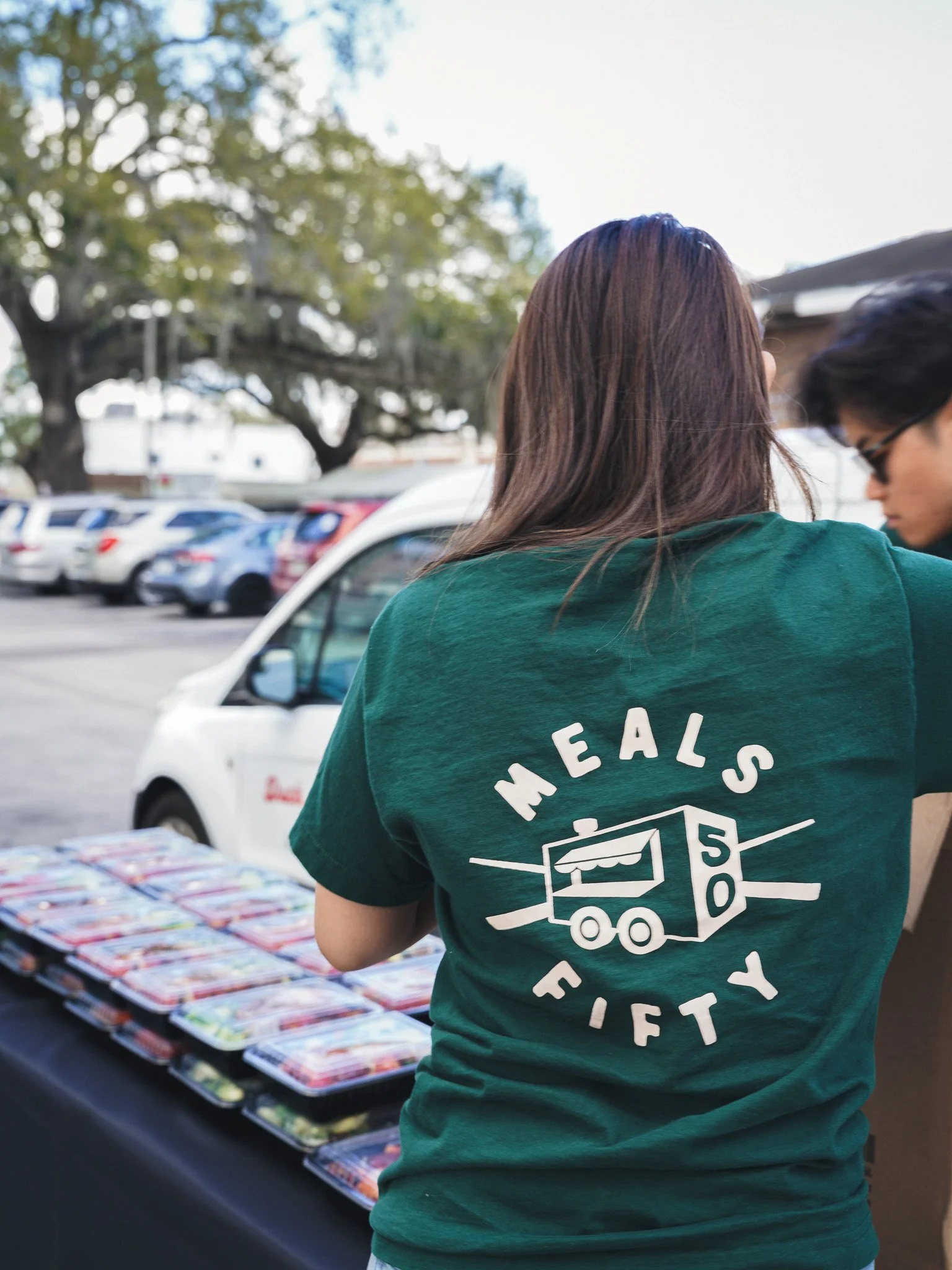 Meals 50 person in a green T-shirt stands near a table with packaged Kai Kai BBQ meals at an outdoor event.