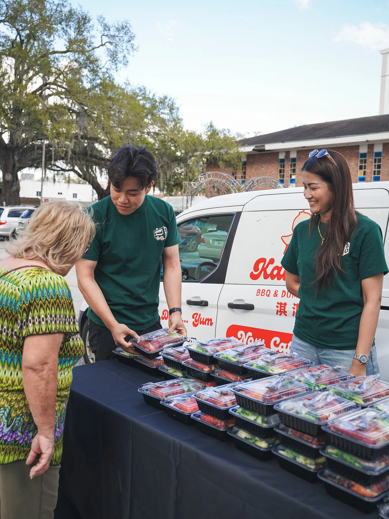 Two young people serve pre-packaged salads to an elderly woman at an outdoor food stall, with a white van and trees with Spanish moss in the background.