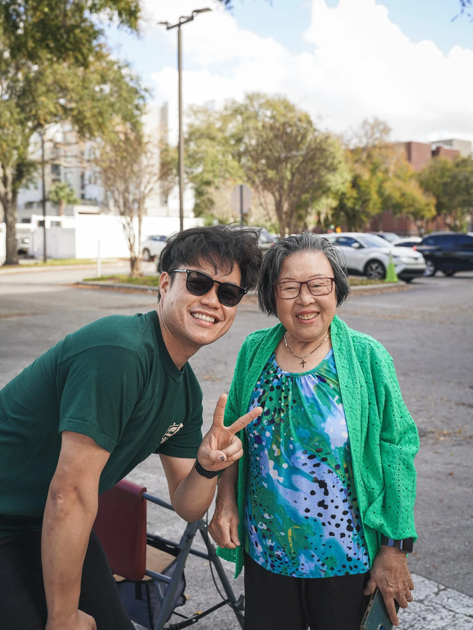 A young man and an elderly woman posing together outdoors on a sunny day, with trees and parked cars in the background. The young man is wearing sunglasses and a green shirt, making a peace sign. The elderly woman is wearing glasses, a colorful dress