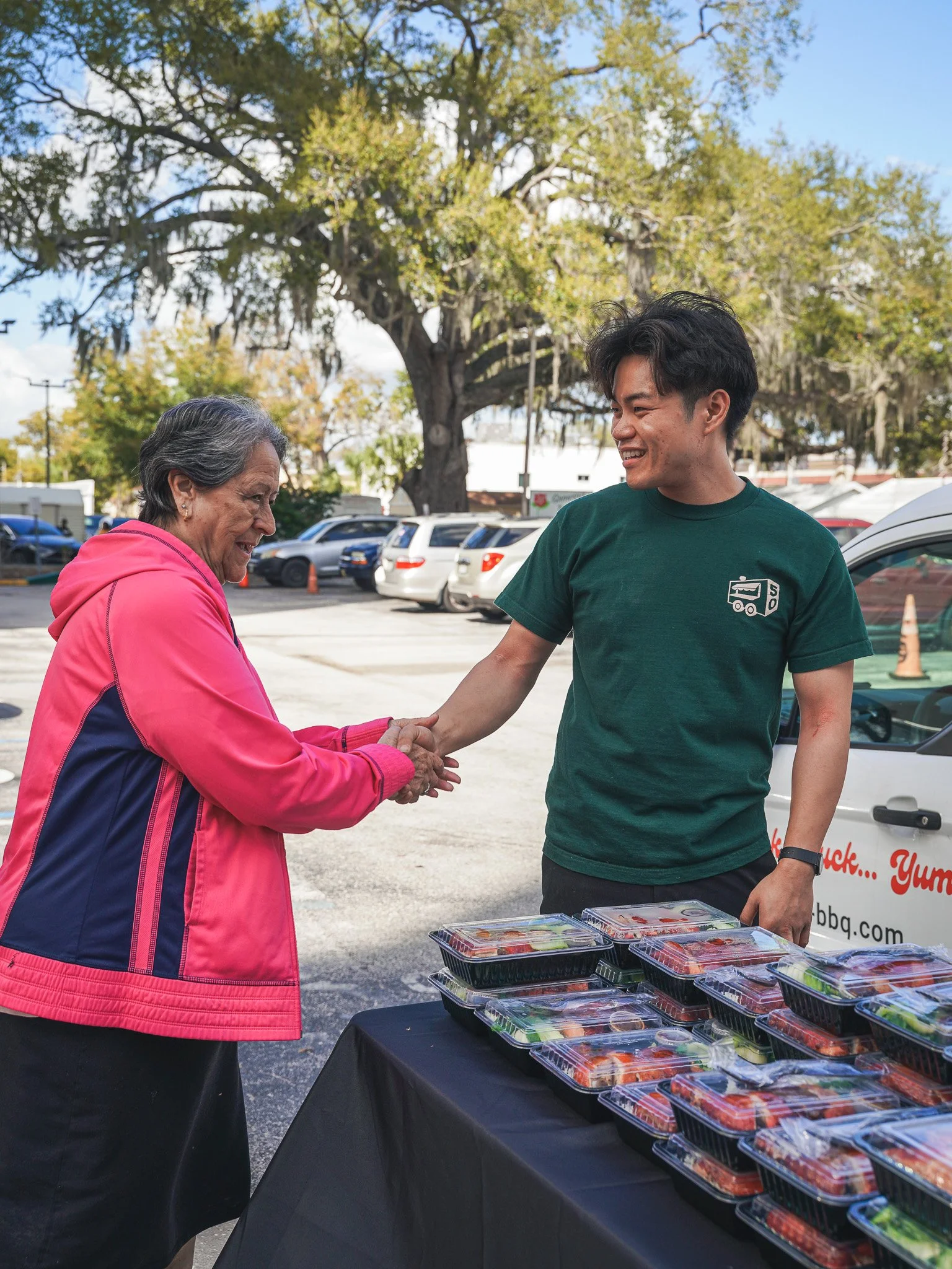 A woman and a young man shake hands at an outdoor table selling ready-to-eat food containers, with a parking lot and trees in the background.