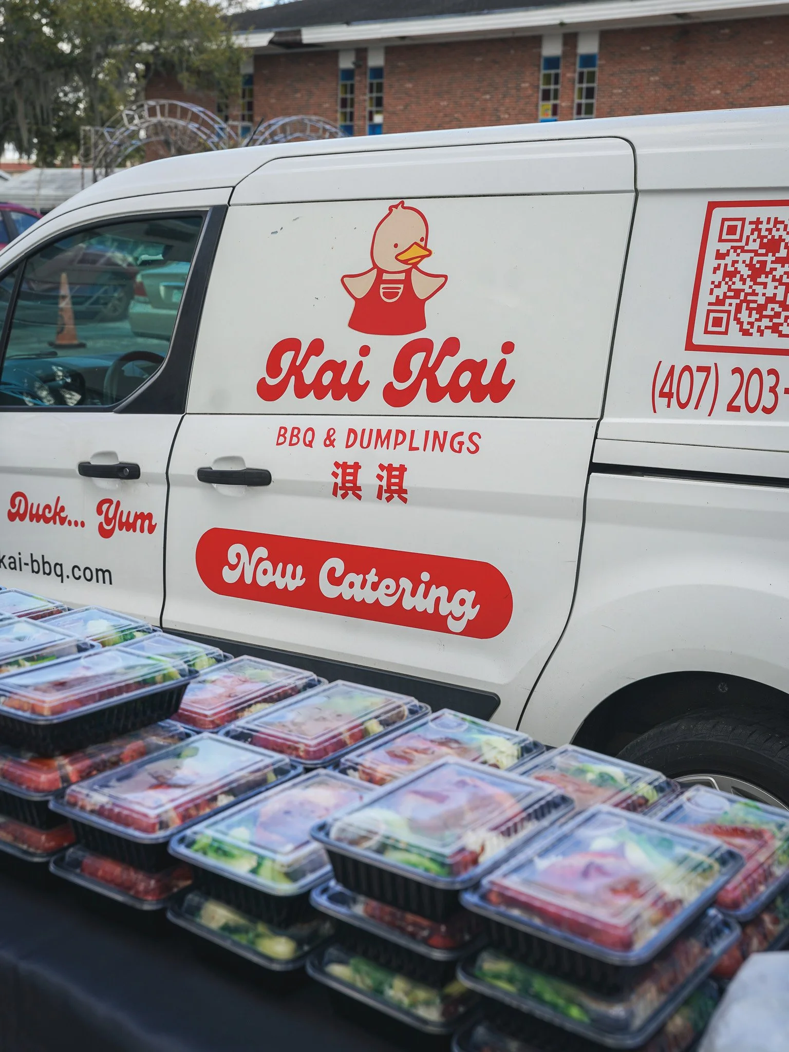 A white delivery van with a red logo of a cartoon duck wearing an apron and the words 'Kai Kai BBQ & Dumplings' on its side, parked behind a table filled with pre-packaged food containers.