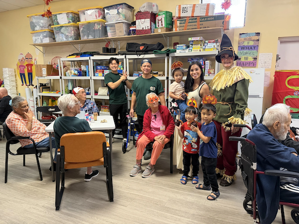 A group of elderly people and children in a room with shelves of supplies, some dressed as scarecrows, posing for a photo.