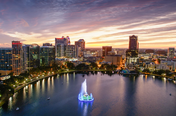 City skyline at sunset with buildings and a lake in the foreground featuring a lit fountain.