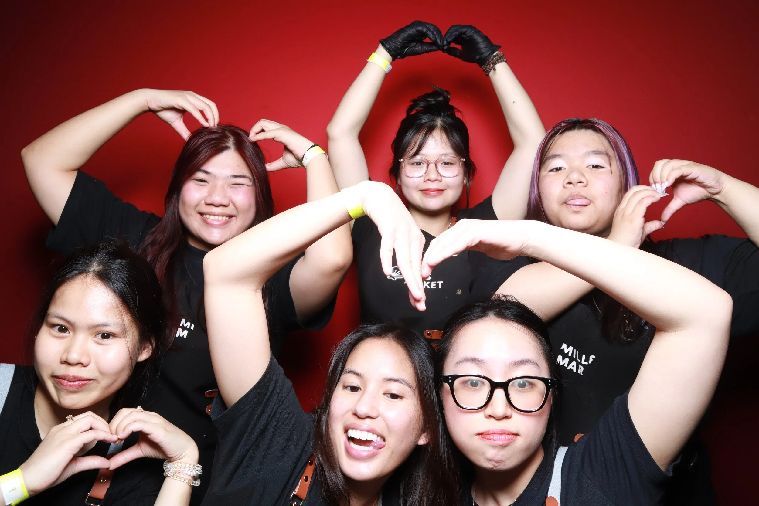 A group of young women taking a selfie against a red background, making heart shapes with their hands.