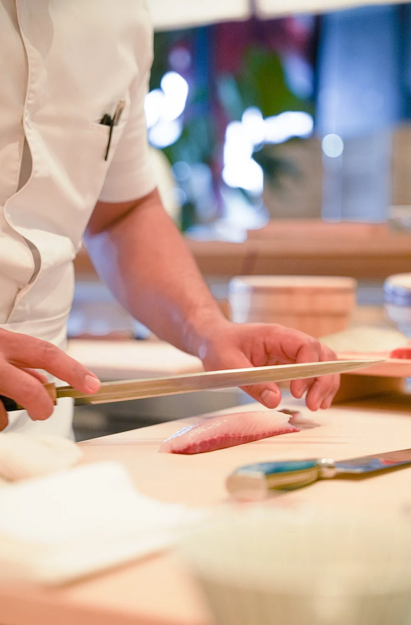 A Okonomi chef preparing sushi, slicing fish on a wooden cutting board in a restaurant kitchen.