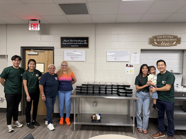 Group of six people, including three young adults, two middle-aged women, and a young girl, standing in a room with a stack of food containers on a metal table, a closed window, and signs on the wall, some wearing green shirts with a logo.