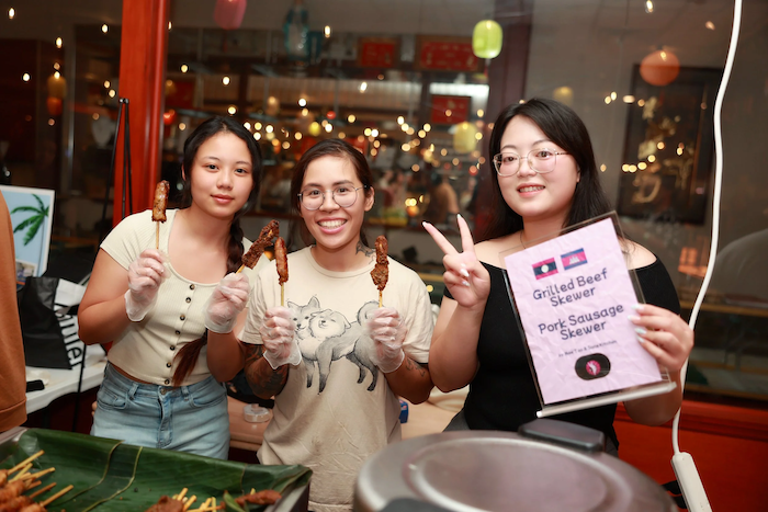 Three women standing behind a food stand, holding grilled meat skewers and smiling, with a sign listing different skewers; one woman making a peace sign with her fingers.