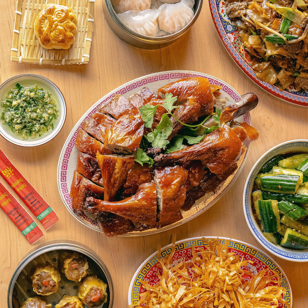 Assorted Kai Kai BBQ dishes on a wooden table, including a roasted chicken, dumplings, stir-fried vegetables, cucumber salad, and sweet dumplings.
