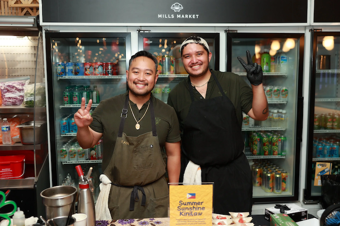 Two smiling men in aprons posing in front of refrigerated drinks at Mills Market.