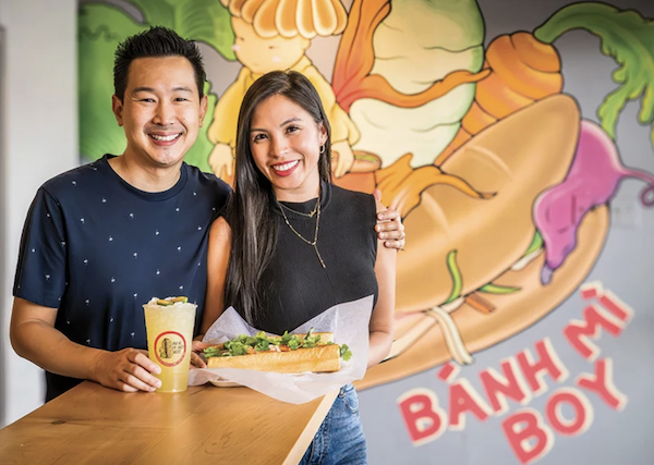 A smiling man and woman stand together inside Mills Market holding a sandwich and a drink, with a colorful cartoon vegetable mural in the background.