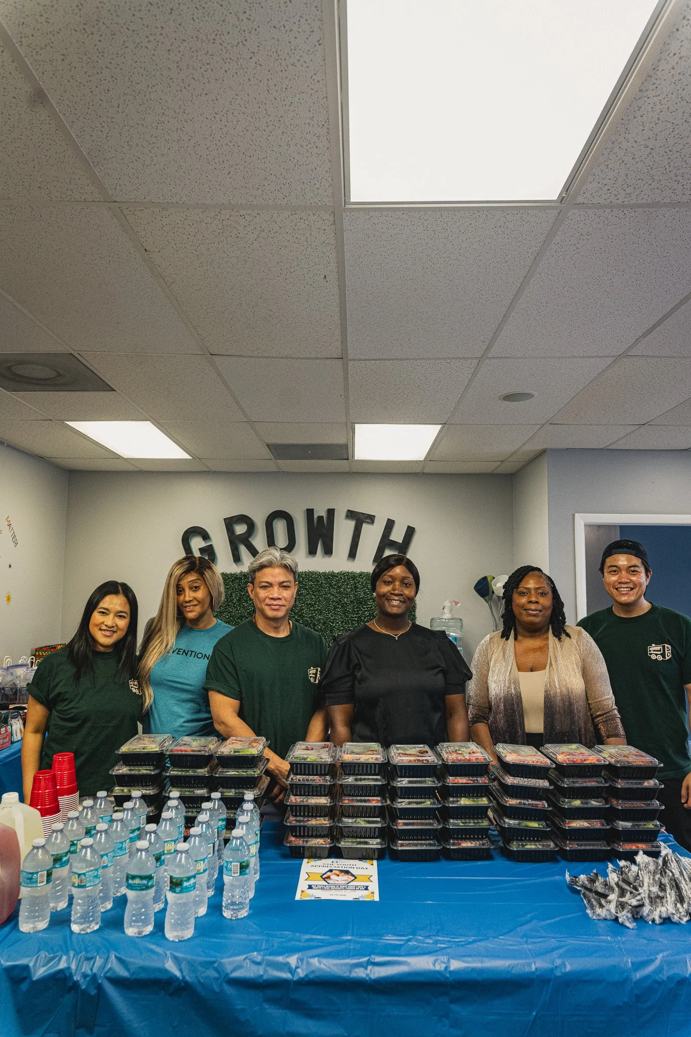 group of 6 smiling team members posing behind a table stacked with hot boxed meals, water bottles, and drinks. 