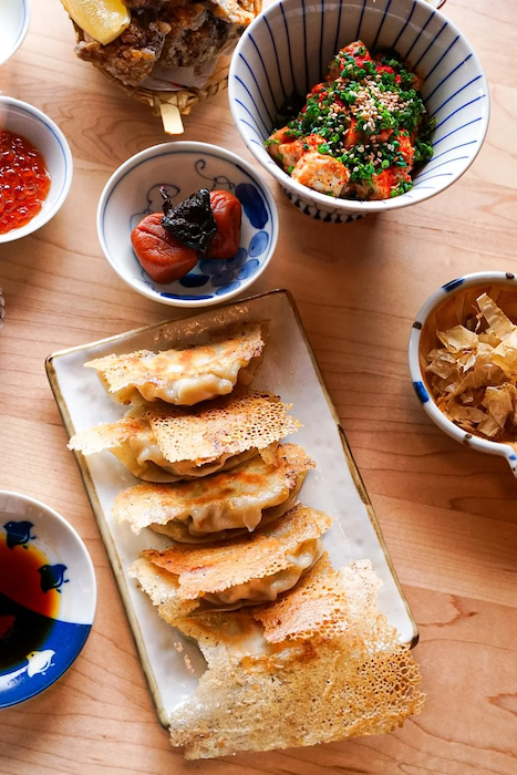 Assorted Zaru dishes on a wooden table, including fried dumplings, pickled vegetables, seaweed in sesame seeds, and small bowls of sauces and condiments.