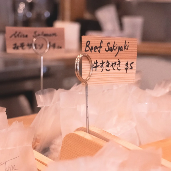 A wooden sign with handwritten text reading 'Beef Sukiyaki $5' and a metal ring stand, placed among plastic-wrapped items at Unigirl.