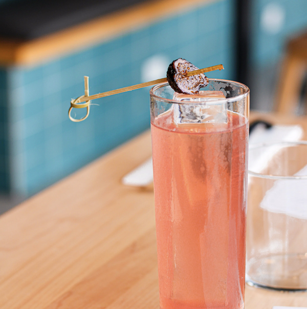 A Zaru pink cocktail, garnished with a slice of grilled pineapple on a skewer, on a wooden table.