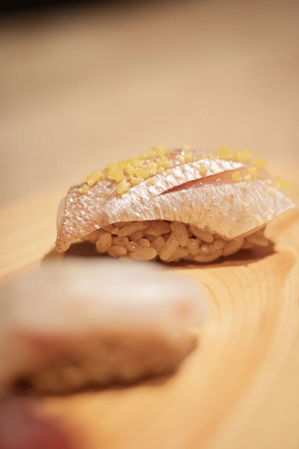 Close-up of a piece of Okonomi sushi with rice, topped with a slice of fish, garnished with yellow grated ginger, on a wooden surface.