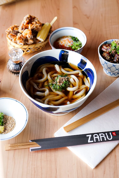 Zaru meal with a bowl of udon noodles, tempura with dipping sauce, and small sides of sauce and garnishes on a wooden table.