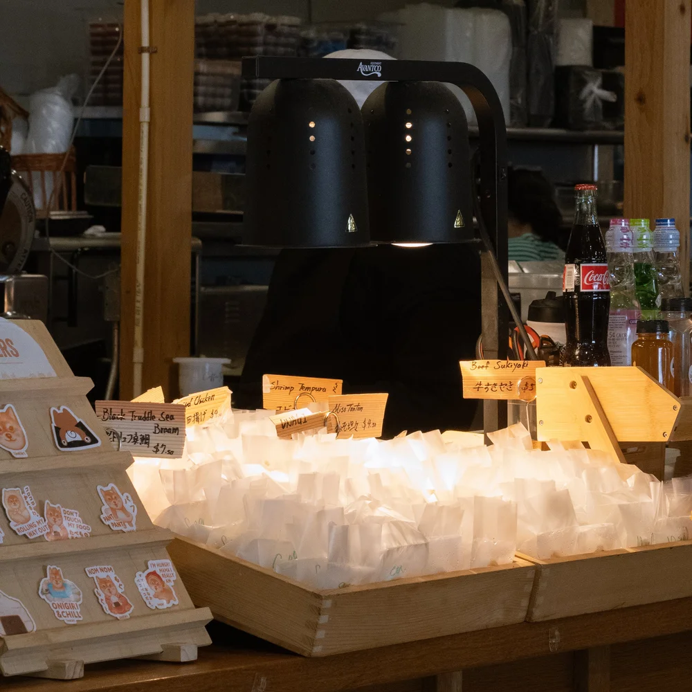 Unigirl shop with a black heat lamp illuminating small paper bags, with handwritten labels including shrimp tempura, beef sukoyaki, and black truffle seaweed bread, and bottled soft drinks in the background.