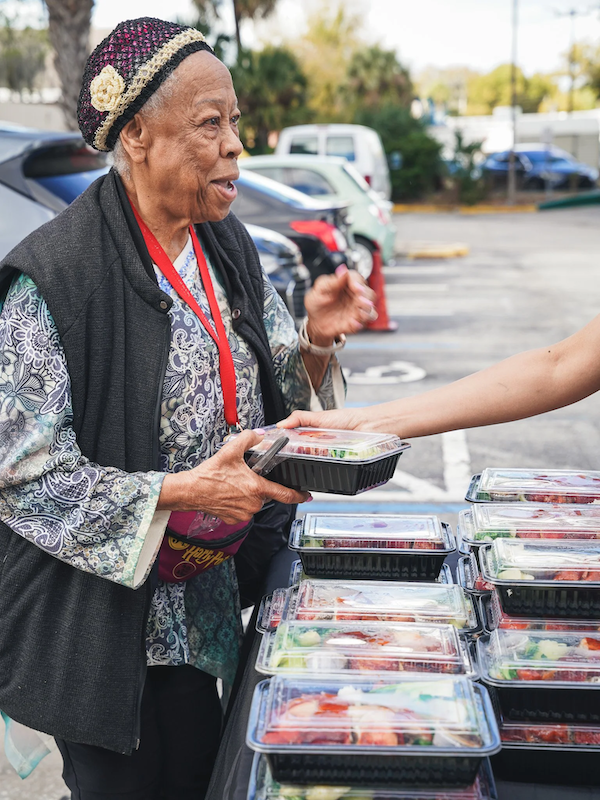 An elderly woman with gray hair, wearing a patterned blouse, black vest, and a colorful knit hat, is buying or selling pre-packaged salads at a Meals 50 events.