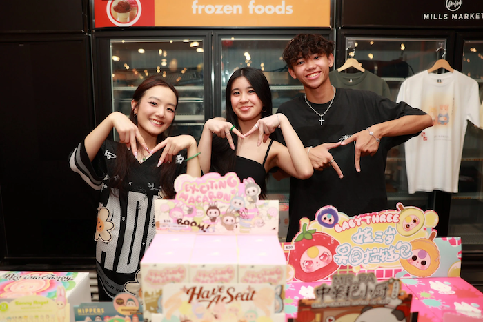 Three young people smiling and posing behind a display table with colorful cartoon-themed decorations and signs, in front of a frozen foods section.