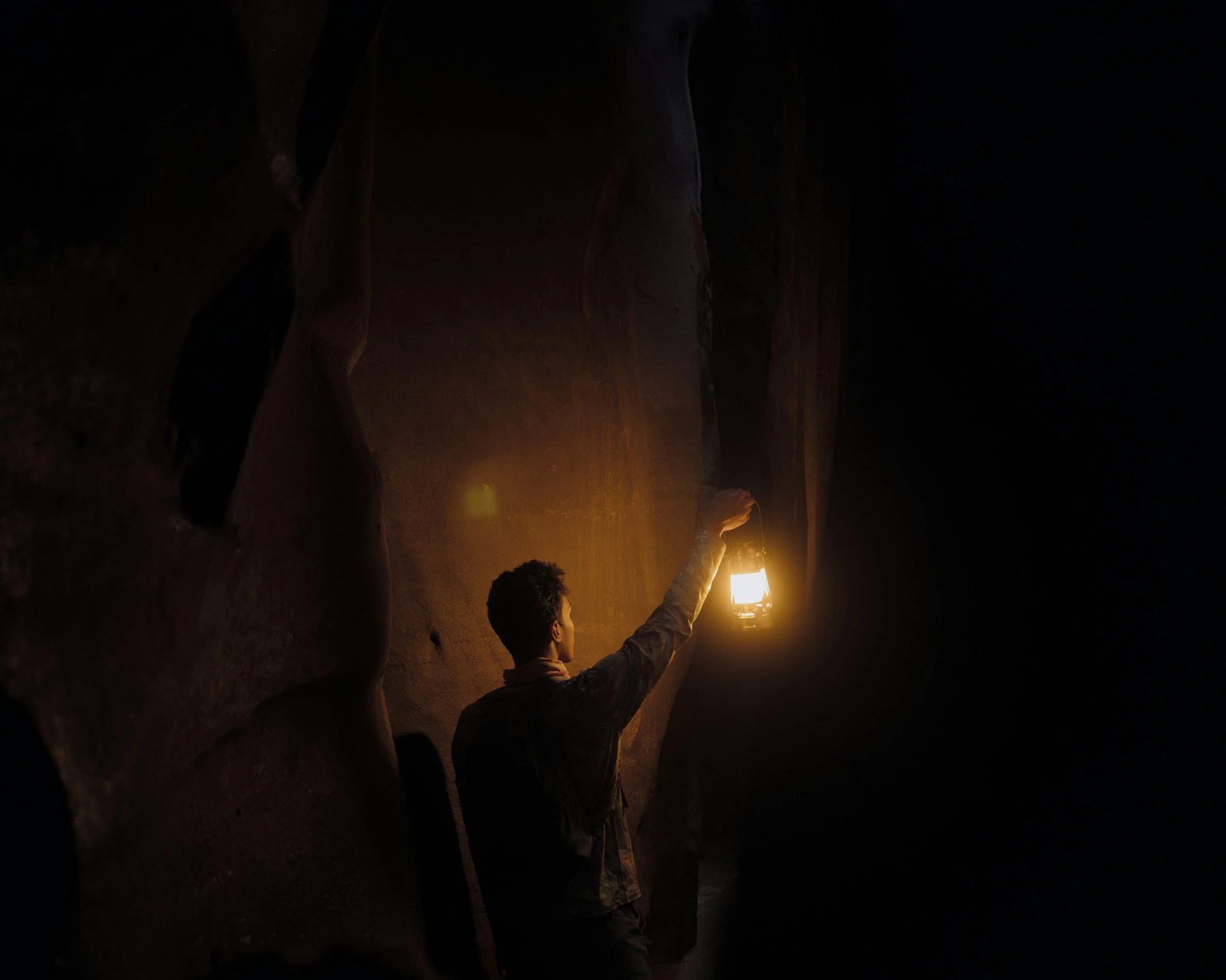 Person holding lantern inside a dark, narrow canyon or cave with high rock walls.