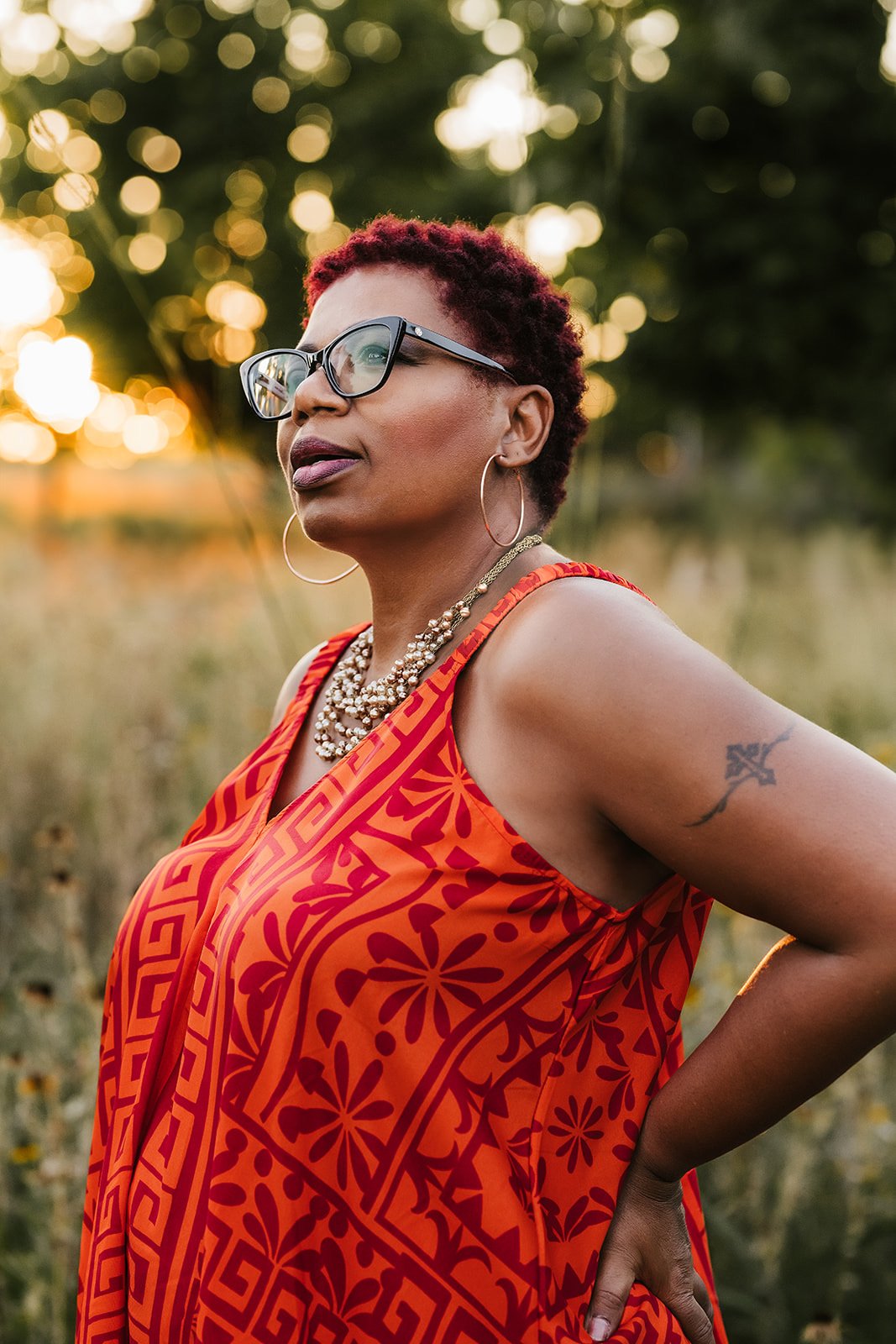 An African American woman with short red hair, wearing large glasses, gold hoop earrings, and a stylish orange dress with red patterns, standing outdoors during sunset.