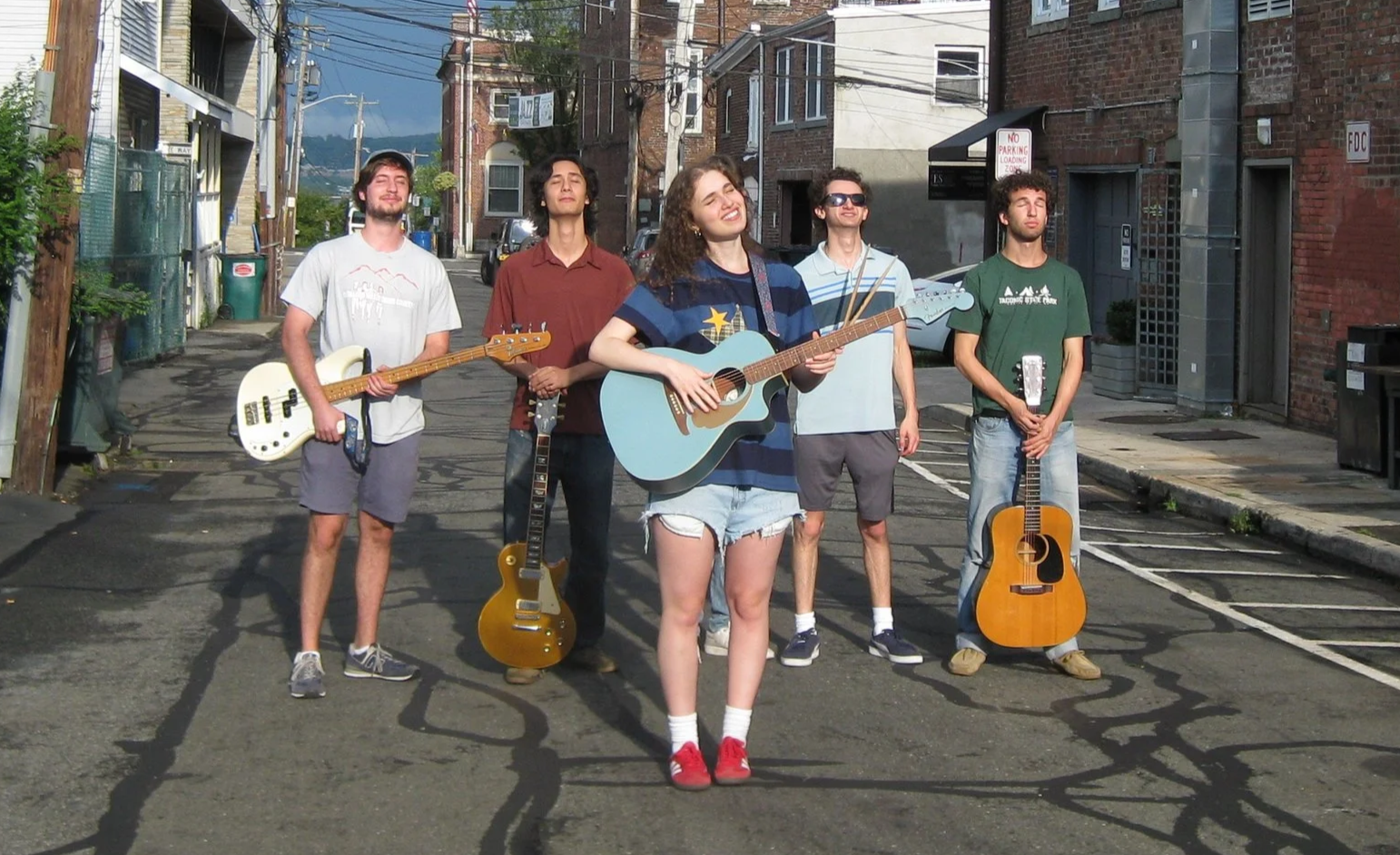 Group of five young musicians standing in an urban alley, holding guitars, with one girl playing an acoustic guitar and smiling, and the others holding electric and acoustic guitars, under a clear sky.
