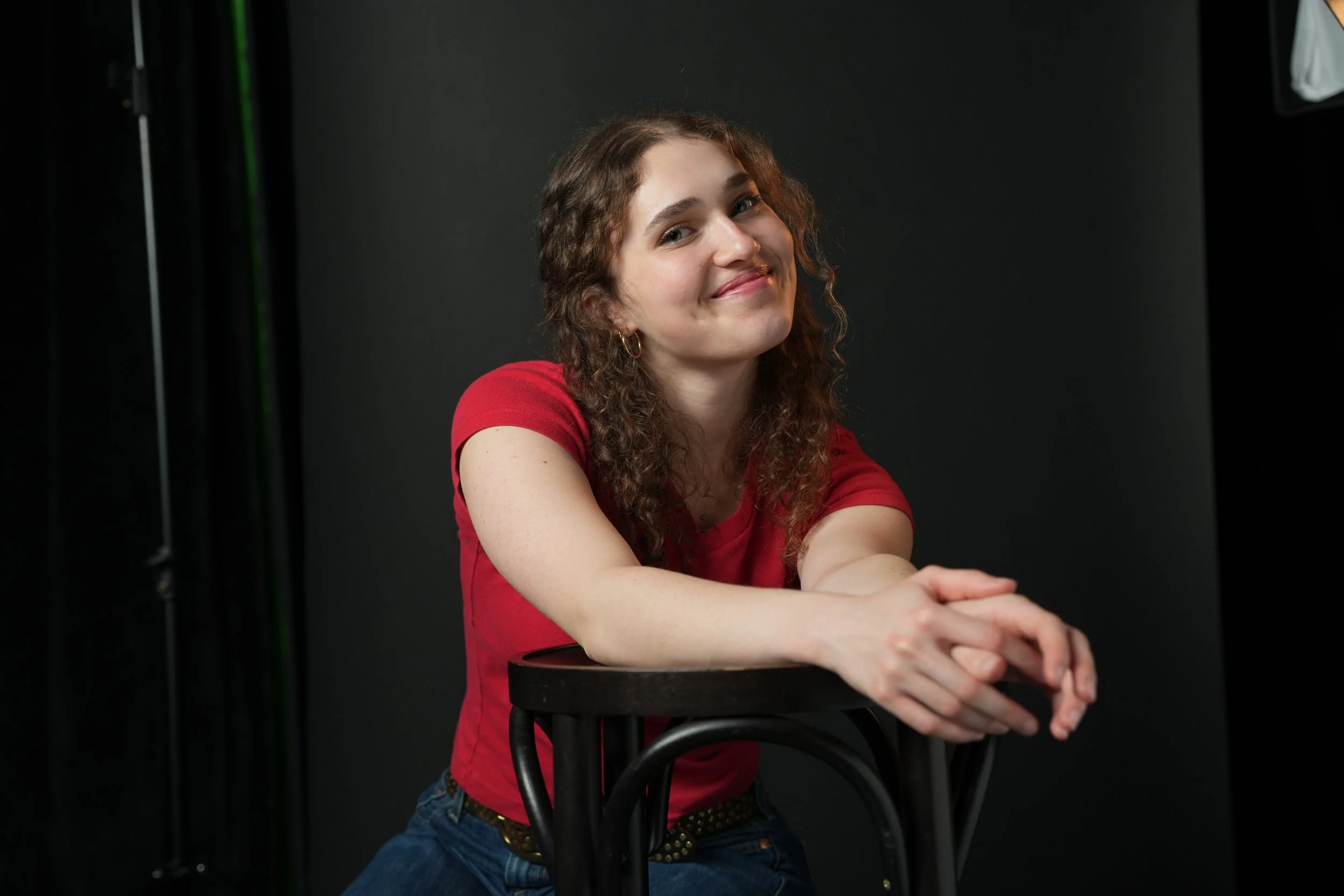 A young woman with curly brown hair and a smile, wearing a red shirt, is sitting at a black stool against a black background.