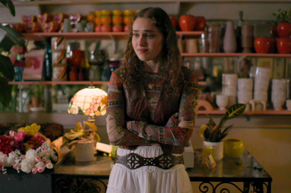 A young woman with curly hair standing in a flower shop, with crossed arms, looking to the side, surrounded by colorful flowers, vases, and decorative items.