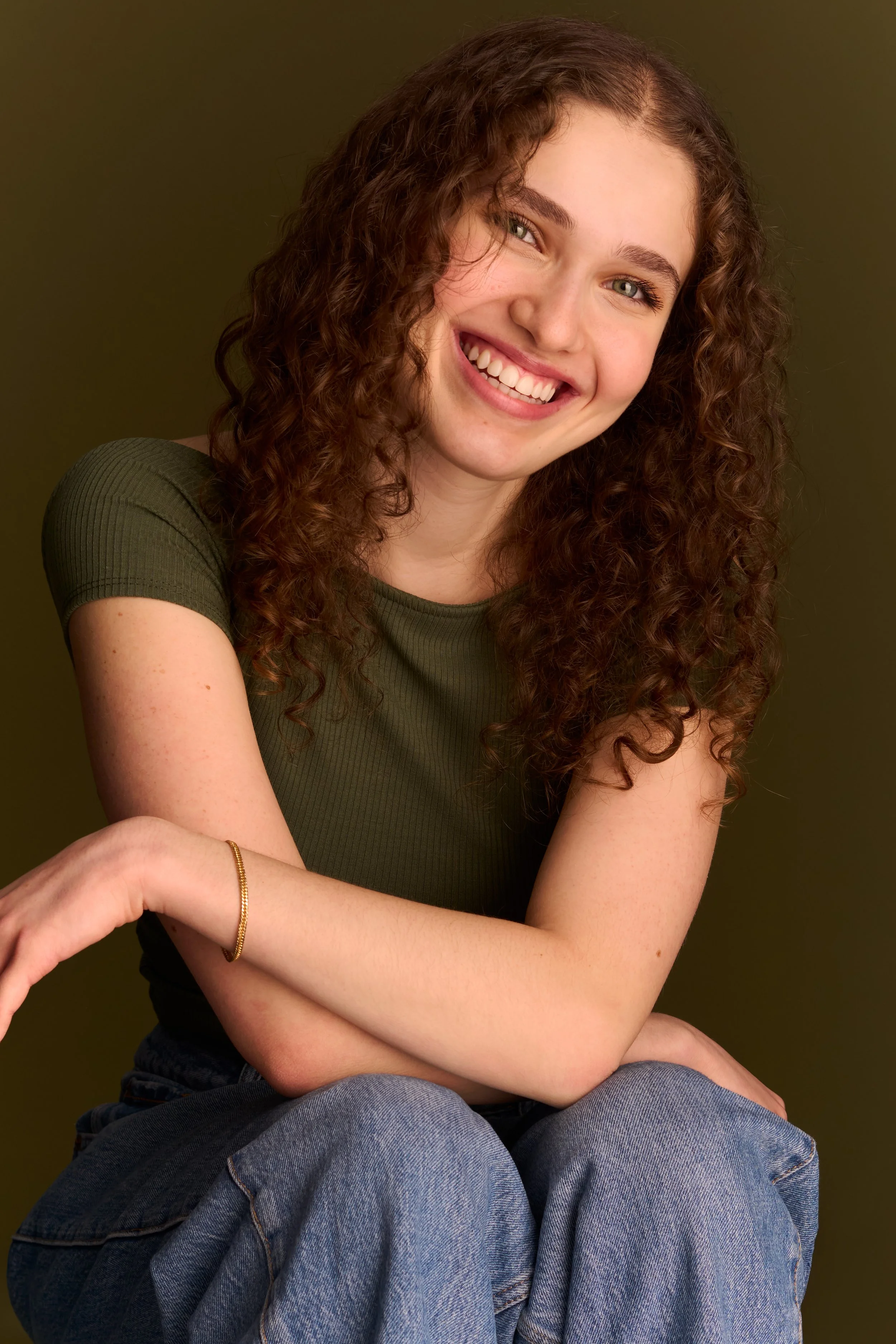A young woman with curly brown hair and green eyes smiling, wearing a green t-shirt and blue jeans, sitting with her arms crossed against a greenish-brown background.