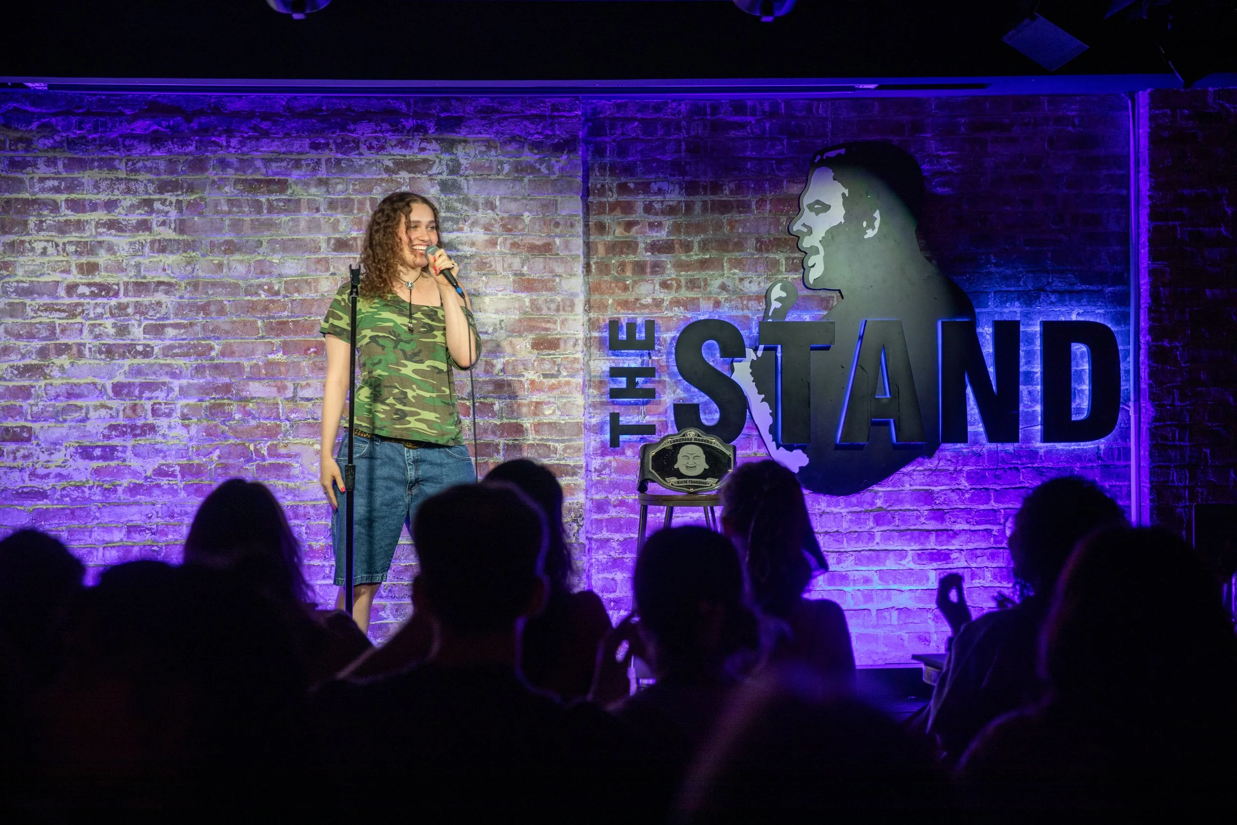 A woman with curly hair wearing a green camouflage t-shirt and jeans is performing stand-up comedy on stage at The Stand comedy club. She is holding a microphone, smiling, and looking toward the audience. The stage has a brick wall background with purple lighting and a large artwork of a woman's face and the words "THE STAND" on the wall.