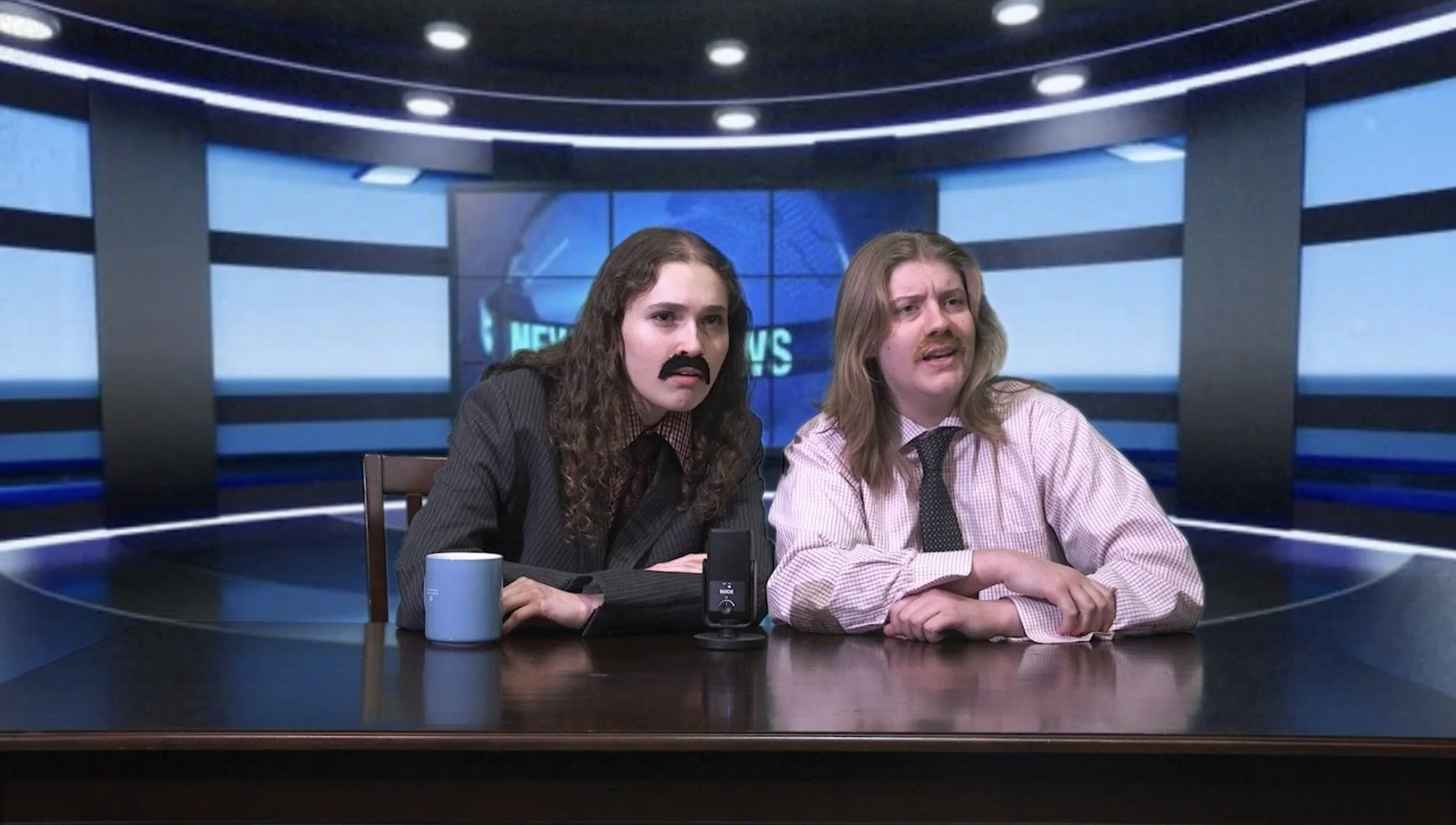 Two men sit at a news desk in a television studio, with one wearing a suit and the other in a shirt and tie, both with long hair and fake mustaches, appearing to be filming a news broadcast