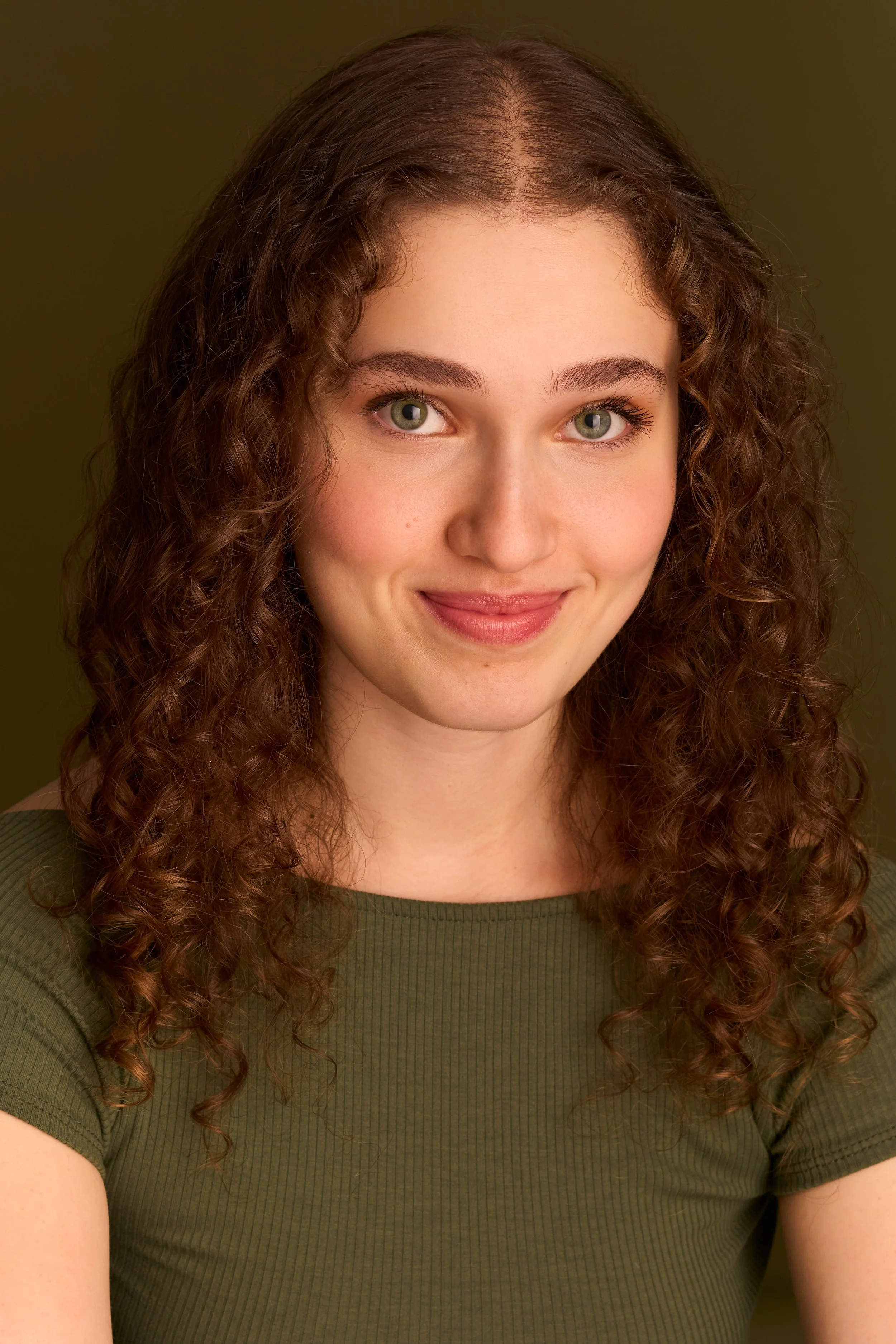 Close-up of a young woman with long, curly brown hair, green eyes, and a slight smile, wearing a green ribbed t-shirt against an olive green background.