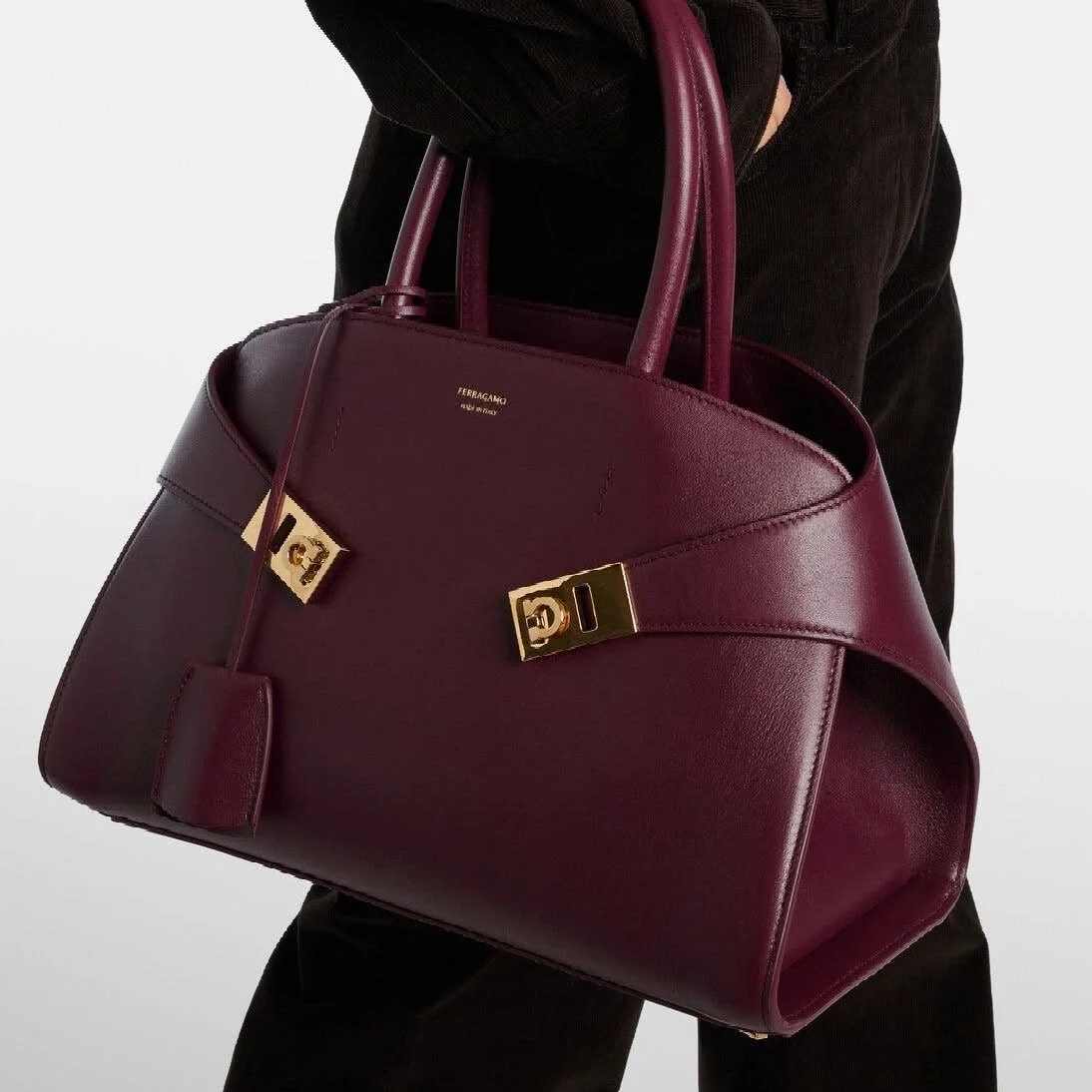 A person holding a large, dark red leather handbag with gold hardware and a small matching luggage tag, against a plain background.