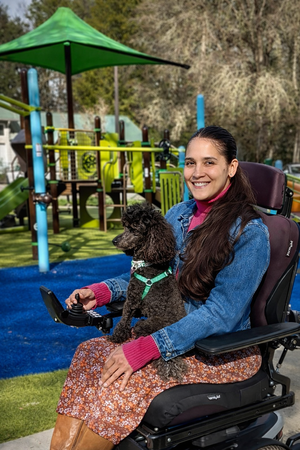 Cameo Ariel K. Perrells, Project Coordinator for the Social Advocacy Network of Disabilities (SAND), smiling while seated in a power wheelchair with her service dog, Minnie, at an accessible community park.