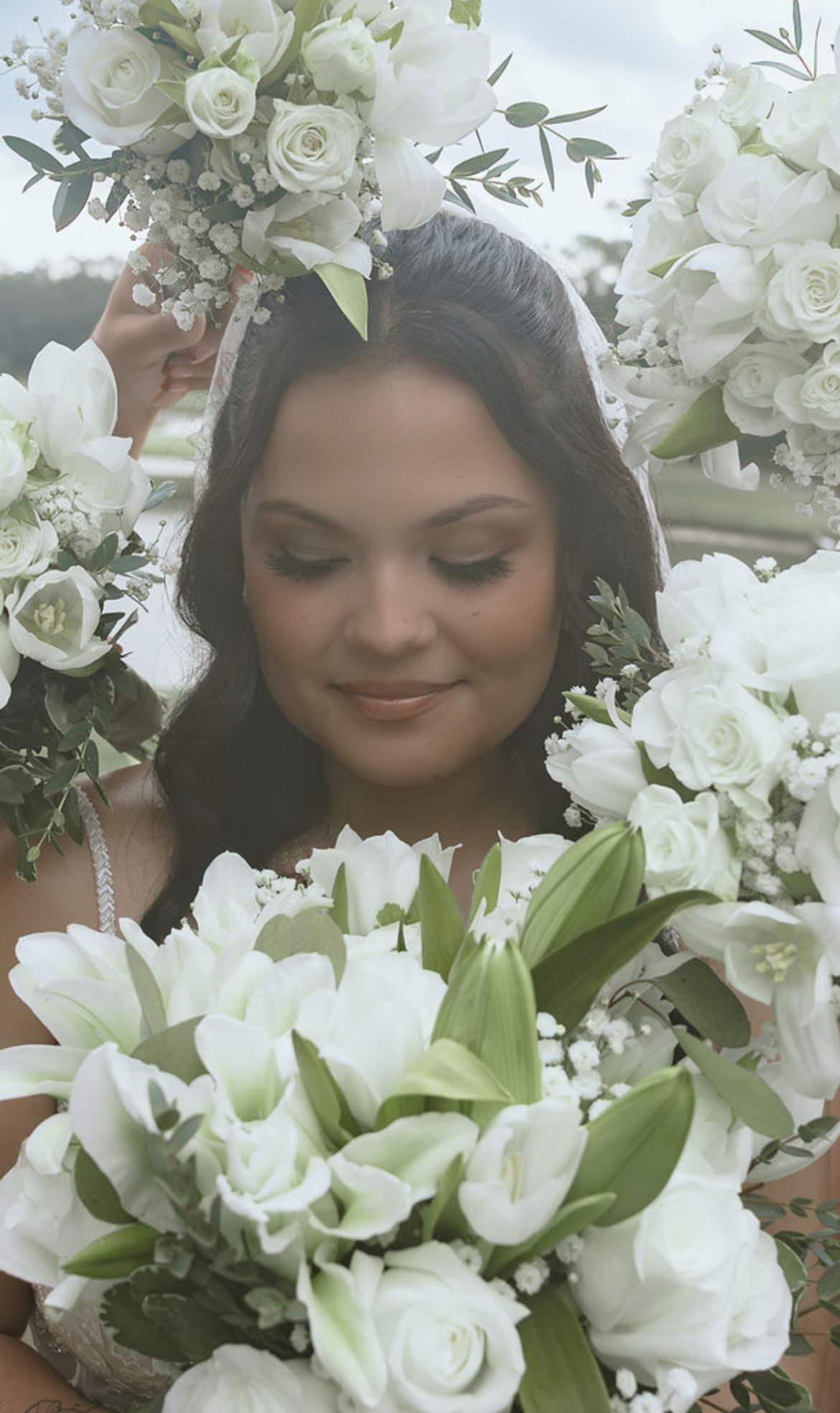 A woman with dark hair and closed eyes, surrounded by white flowers including roses and lilies, holding a large bouquet of flowers above her head.