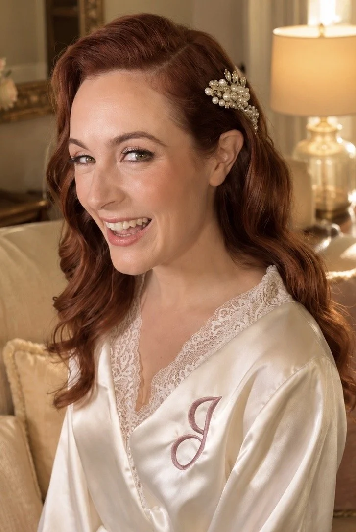 A smiling woman with red hair styled in loose waves, wearing a cream-colored satin robe with lace trim and a pearl hair accessory, sitting in a warmly lit room.