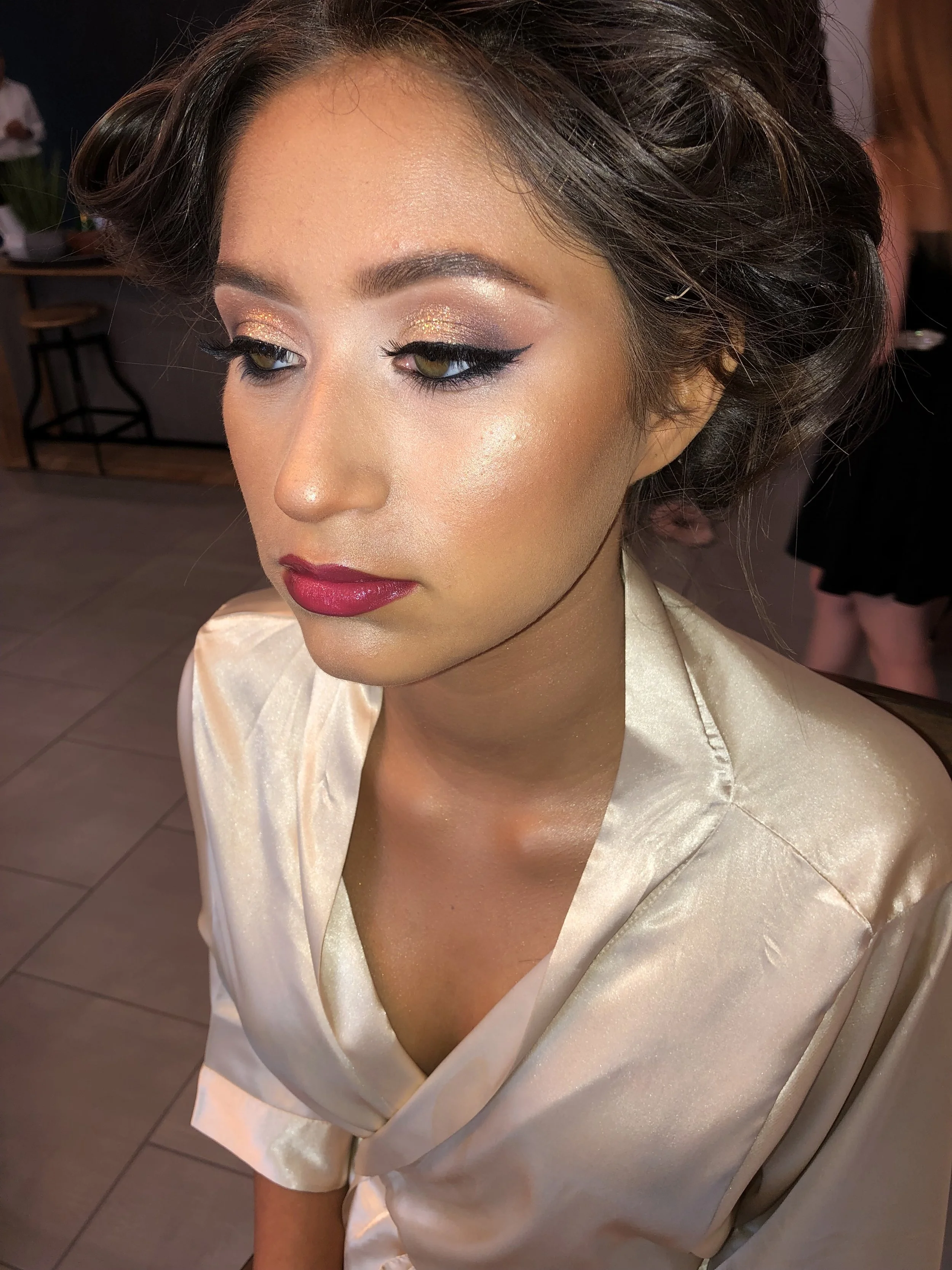 Close-up of a woman with styled brown hair, makeup including gold and pink eyeshadow, winged eyeliner, false eyelashes, and red lipstick, wearing a satin cream-colored robe.