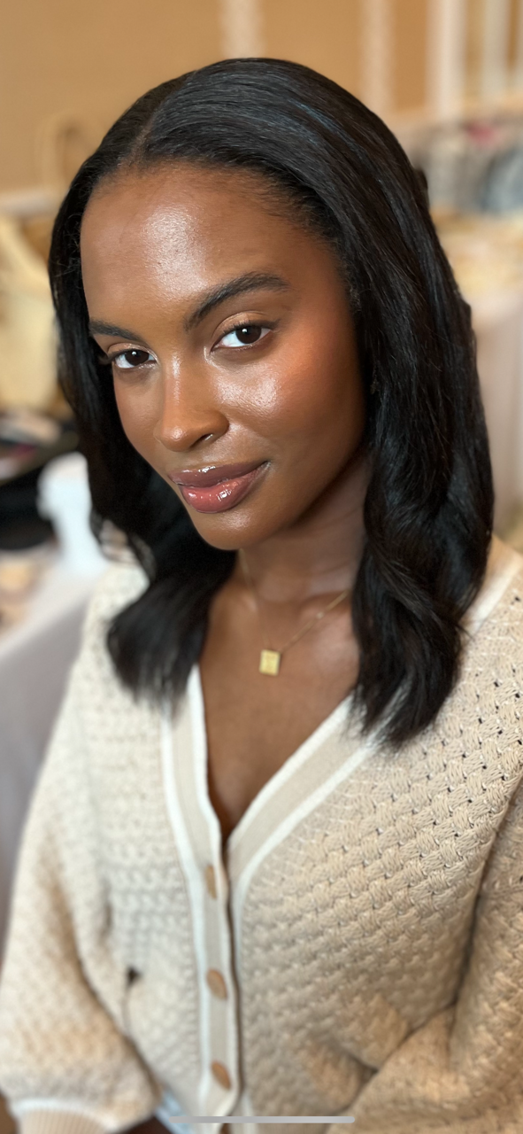 A woman with black hair and brown skin, wearing a cream-colored sweater, gold necklace, and natural makeup, smiling slightly, indoors.
