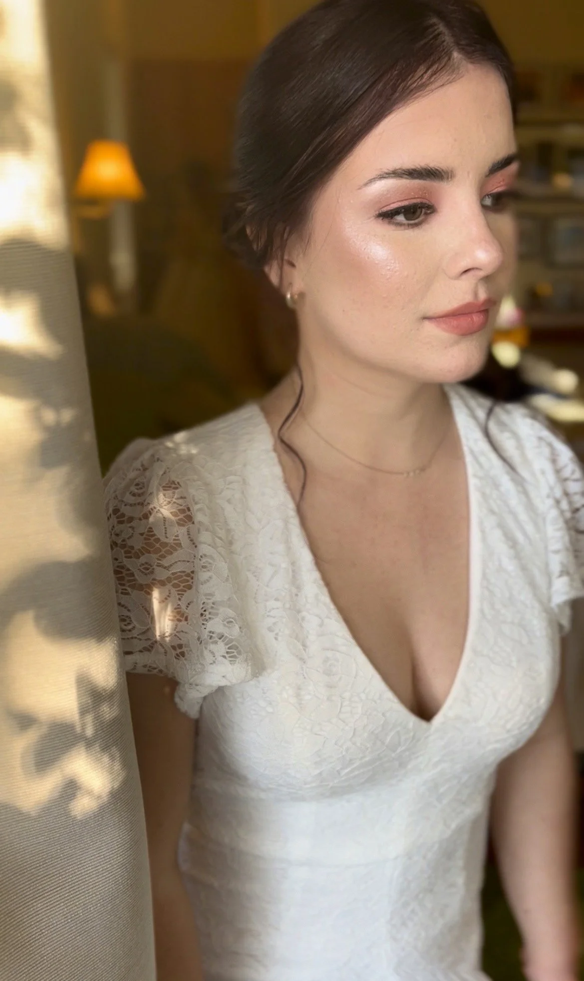 A woman with long brown hair styled in loose waves, wearing a white lace dress with short sleeves and a V-neckline, standing indoors with a warm background including a lamp and shelves.