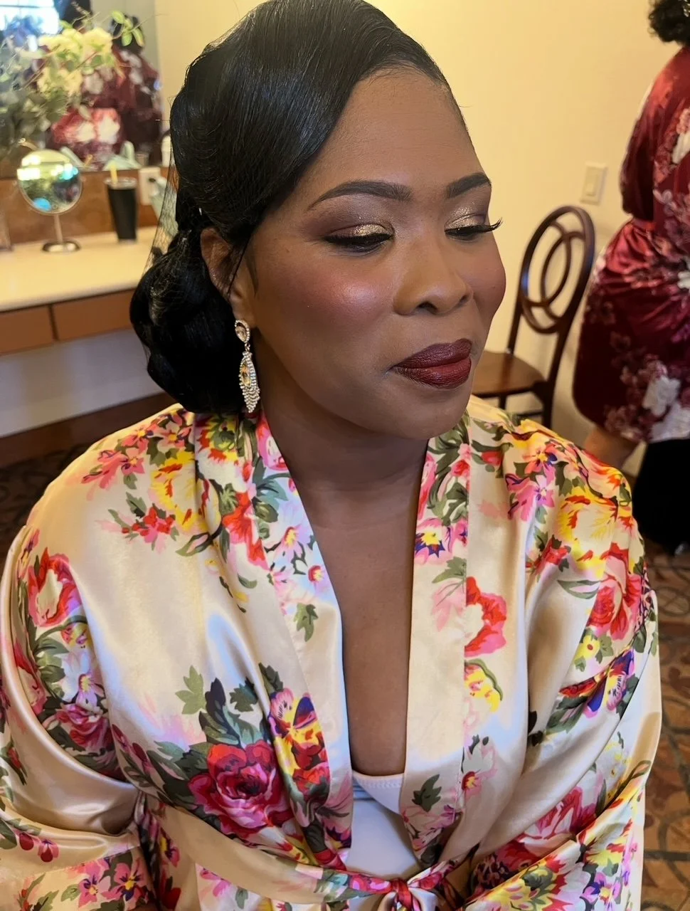 A woman with dark hair styled in waves, wearing makeup with gold eyeshadow and dark lipstick, dressed in a floral satin robe, and wearing dangling earrings. She is seated with her eyes closed, possibly at a special event or occasion.