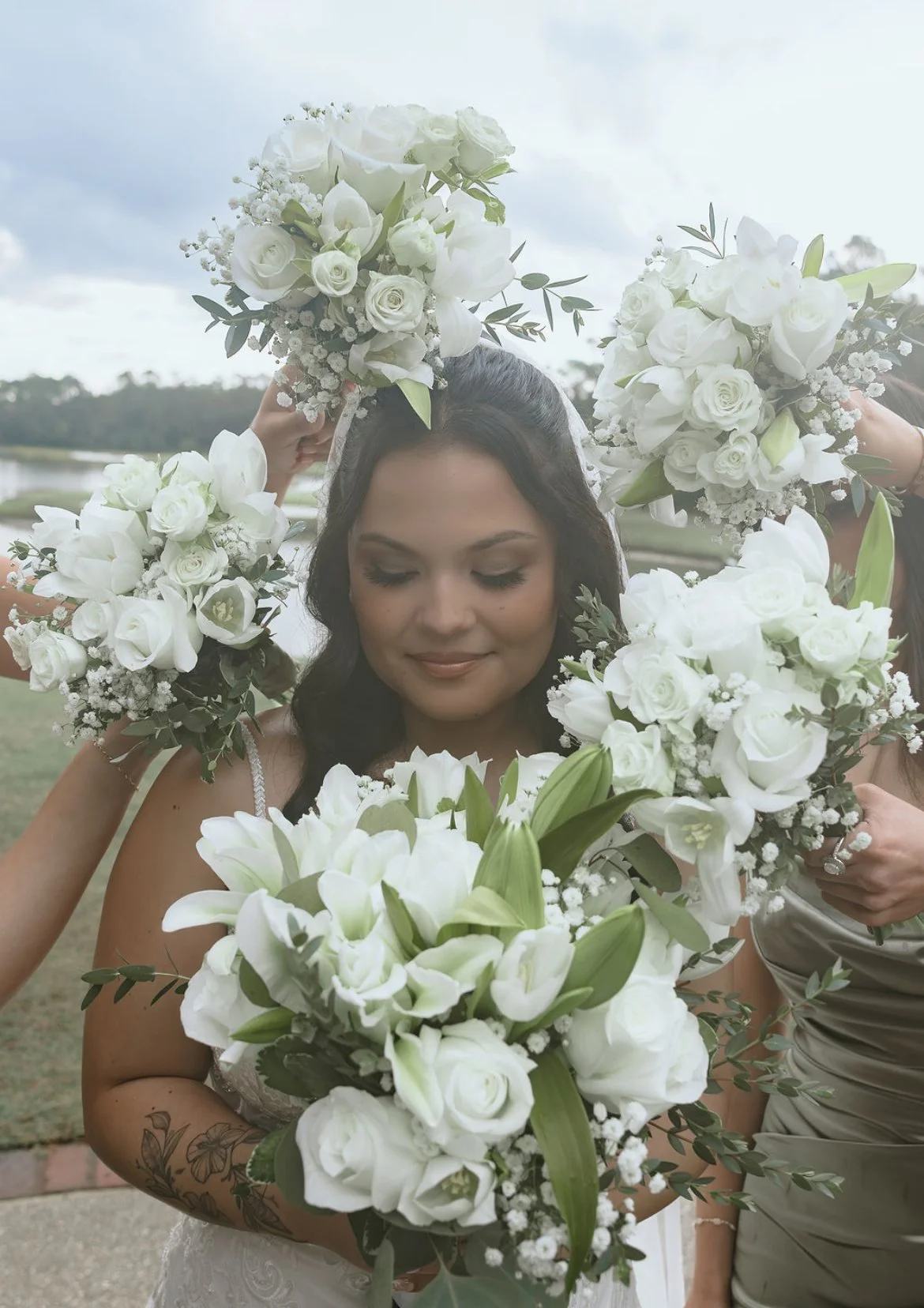 A bride with dark hair and closed eyes holds a bouquet of white lilies and roses, surrounded by friends also holding white floral arrangements, outdoors on a cloudy day.