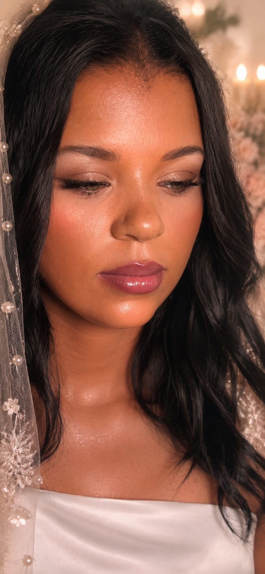 Close-up of a woman with dark hair, wearing makeup, dressed in white, with a sheer veil decorated with pearls and floral embroidery, in a softly lit setting with candles and flowers in the background.